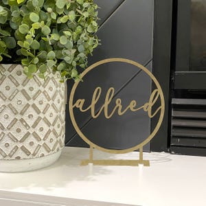 Gold metal tabletop nameplate with \“allred\“ in cursive inside a circle, placed next to a decorative plant in a textured white pot.