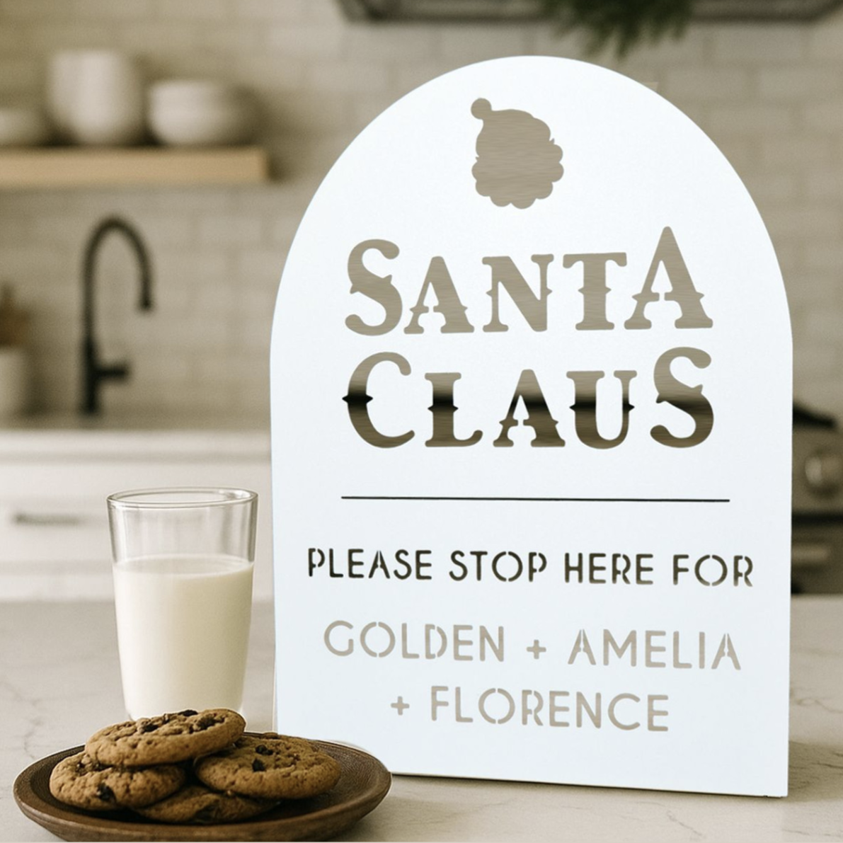 A decorative sign for Santa Claus requests his stop and includes the names Golden, Amelia, and Florence. It is placed on a countertop next to a glass of milk and a plate of cookies.