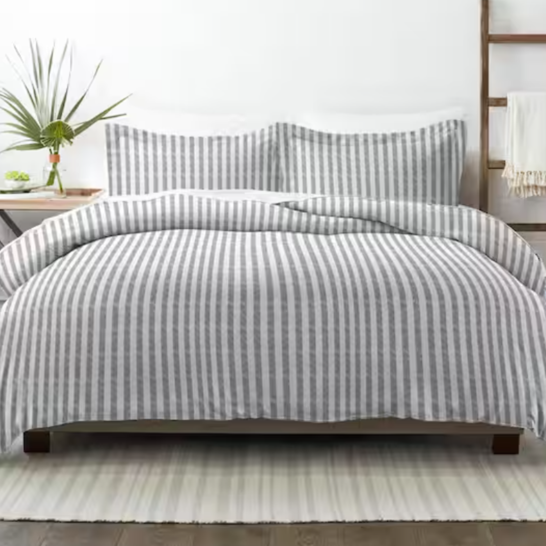 A bed features a gray and white striped comforter and matching pillow shams. A small plant decorates the bedside table, and a white rug lies underneath.