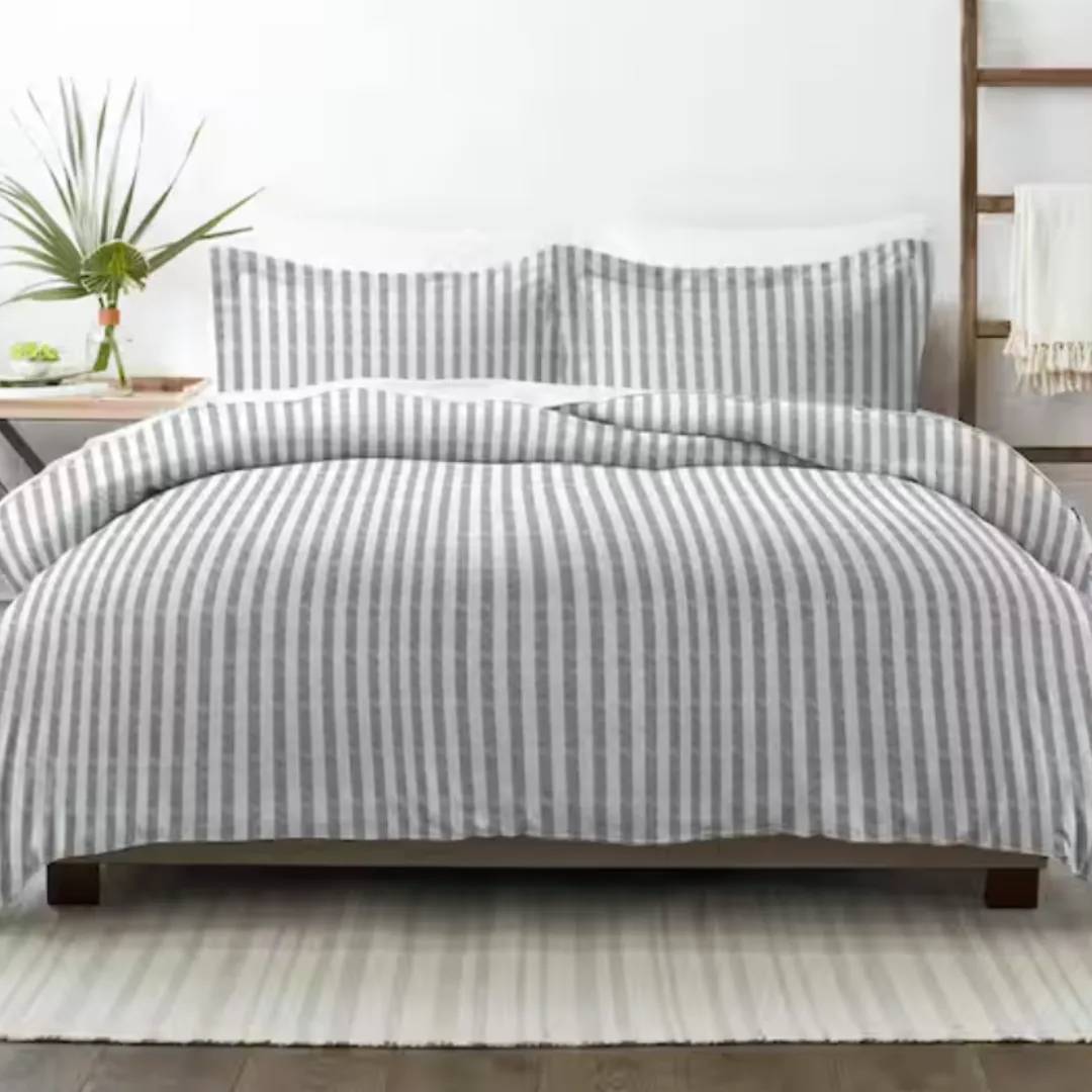A bed features a gray and white striped comforter and matching pillow shams. A small plant decorates the bedside table, and a white rug lies underneath.