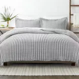 A bed features a gray and white striped comforter and matching pillow shams. A small plant decorates the bedside table, and a white rug lies underneath.