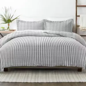 A bed features a gray and white striped comforter and matching pillow shams. A small plant decorates the bedside table, and a white rug lies underneath.