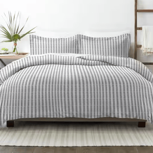 A bed features a gray and white striped comforter and matching pillow shams. A small plant decorates the bedside table, and a white rug lies underneath.