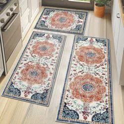 Two decorative kitchen mats with intricate floral patterns in orange, blue, and white are placed on a light wood floor. They are positioned near a stove and sink, adjacent to a window with a potted plant.