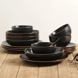 A set of matte black dinnerware, including plates and bowls with a subtle brown rim, is arranged on a wooden table.