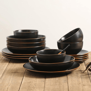 A set of matte black dinnerware, including plates and bowls with a subtle brown rim, is arranged on a wooden table.