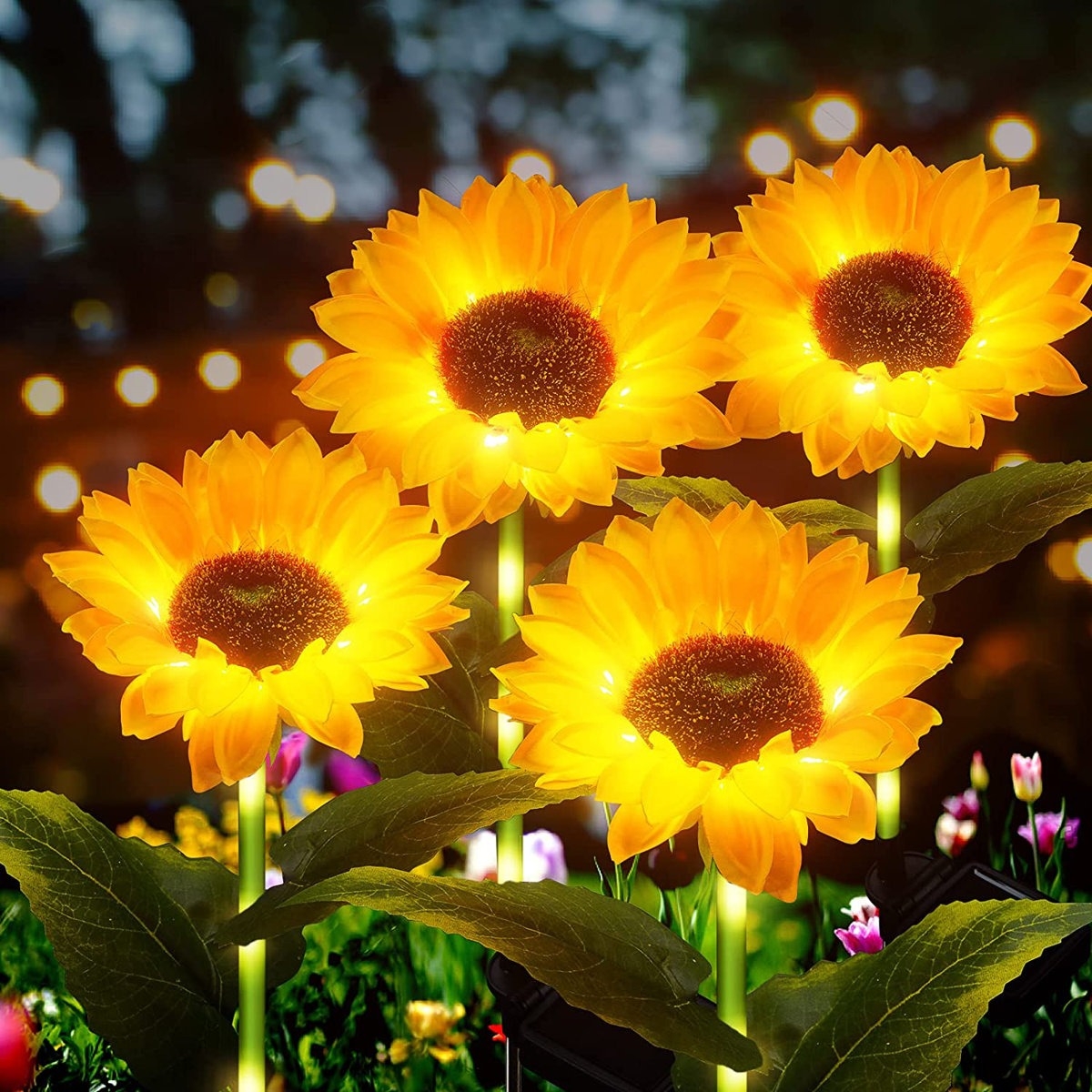 Four LED solar-powered sunflower garden lights are glowing brightly in a nighttime setting, surrounded by other foliage and lights.