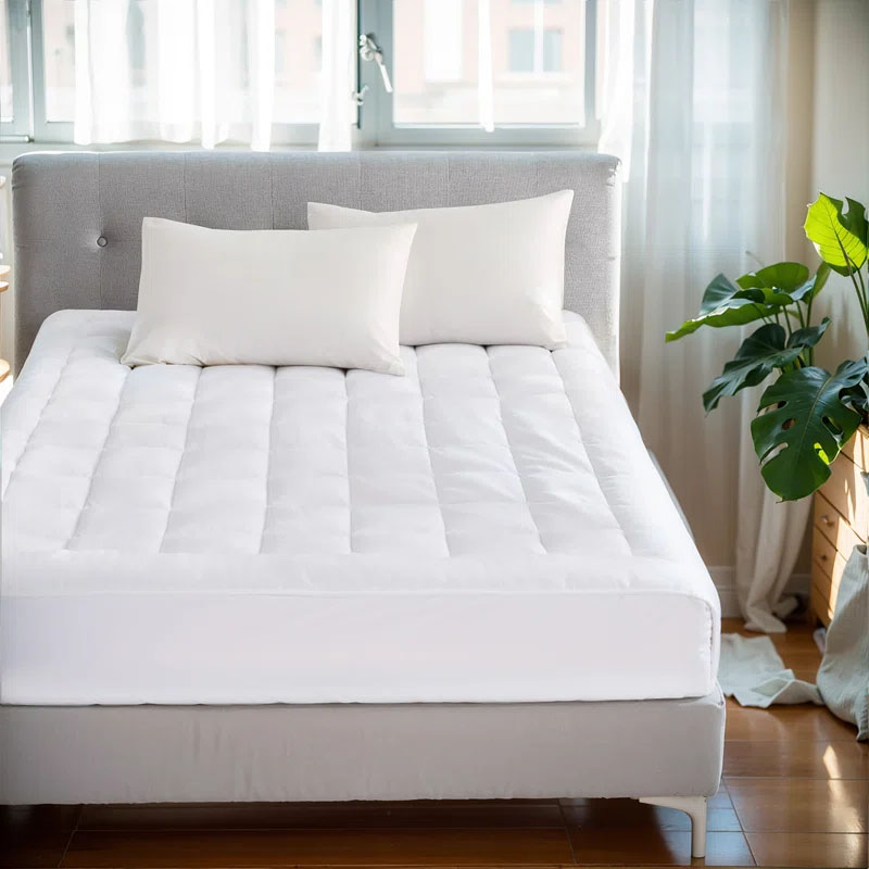 White overfilled waterproof mattress pad on a bed with a quilted design, accompanied by two white pillows. The setting includes a light gray headboard and a green plant beside the bed, located in a sunlit room with sheer curtains.