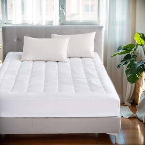 White overfilled waterproof mattress pad on a bed with a quilted design, accompanied by two white pillows. The setting includes a light gray headboard and a green plant beside the bed, located in a sunlit room with sheer curtains.