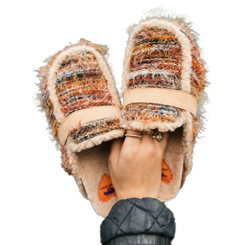 A pair of fuzzy, multicolored slippers with a textured weave and cozy lining are held in a hand.