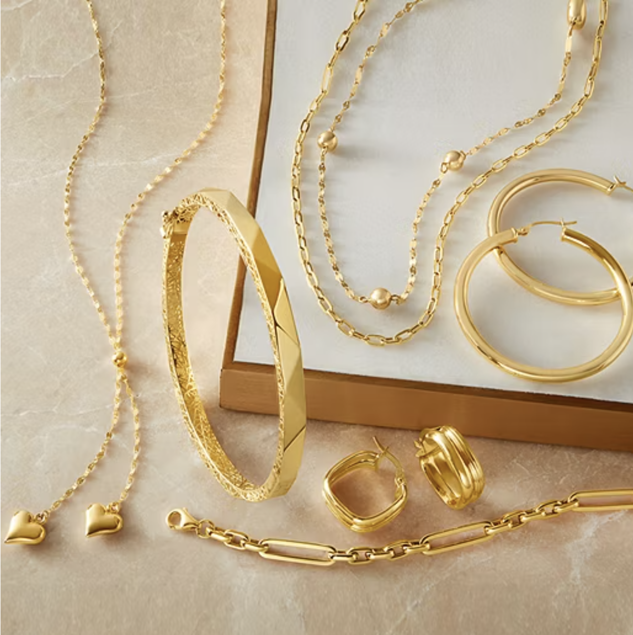 A collection of gold jewelry including necklaces, hoop earrings, a bangle, stud earrings with heart-shaped charms, and a link bracelet arranged on a flat surface.