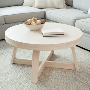 A round, light wood coffee table with a simple, clean design and sturdy crossed legs. It features a bowl with decorative items and two stacked books on top, placed on a light-colored textured rug next to a gray sectional sofa.