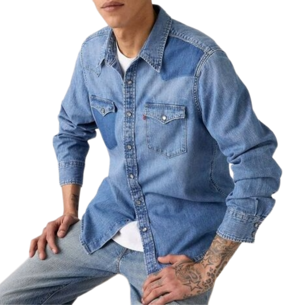 A person with tattoos is wearing a light blue denim shirt with snap-button pockets over a white T-shirt, paired with light blue jeans.