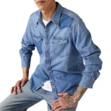 A person with tattoos is wearing a light blue denim shirt with snap-button pockets over a white T-shirt, paired with light blue jeans.