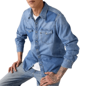 A person with tattoos is wearing a light blue denim shirt with snap-button pockets over a white T-shirt, paired with light blue jeans.