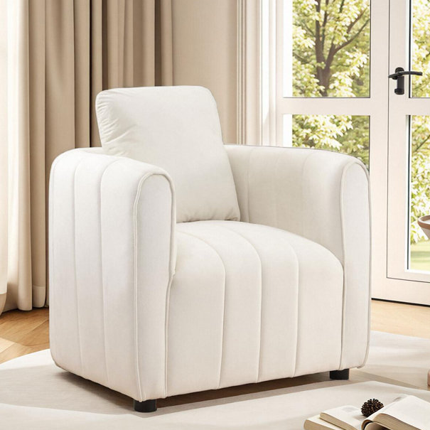 A white upholstered armchair with a modern design is placed in a room with wooden flooring and large windows. The chair features vertical stitching and appears comfortable with plush cushions.