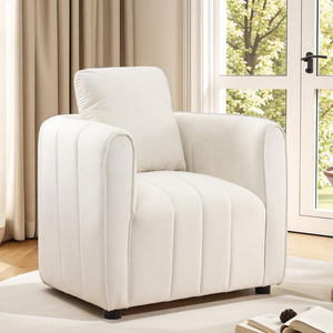 A white upholstered armchair with a modern design is placed in a room with wooden flooring and large windows. The chair features vertical stitching and appears comfortable with plush cushions.