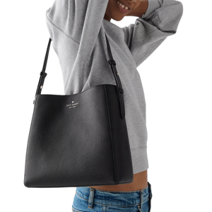 A person carries a black Kate Spade New York tote bag with adjustable straps, wearing a gray sweatshirt and blue jeans.