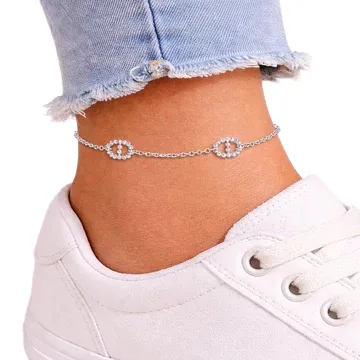 A delicate silver anklet with circular diamond-like accents, worn above a pair of light wash, frayed hem jeans and white sneakers.