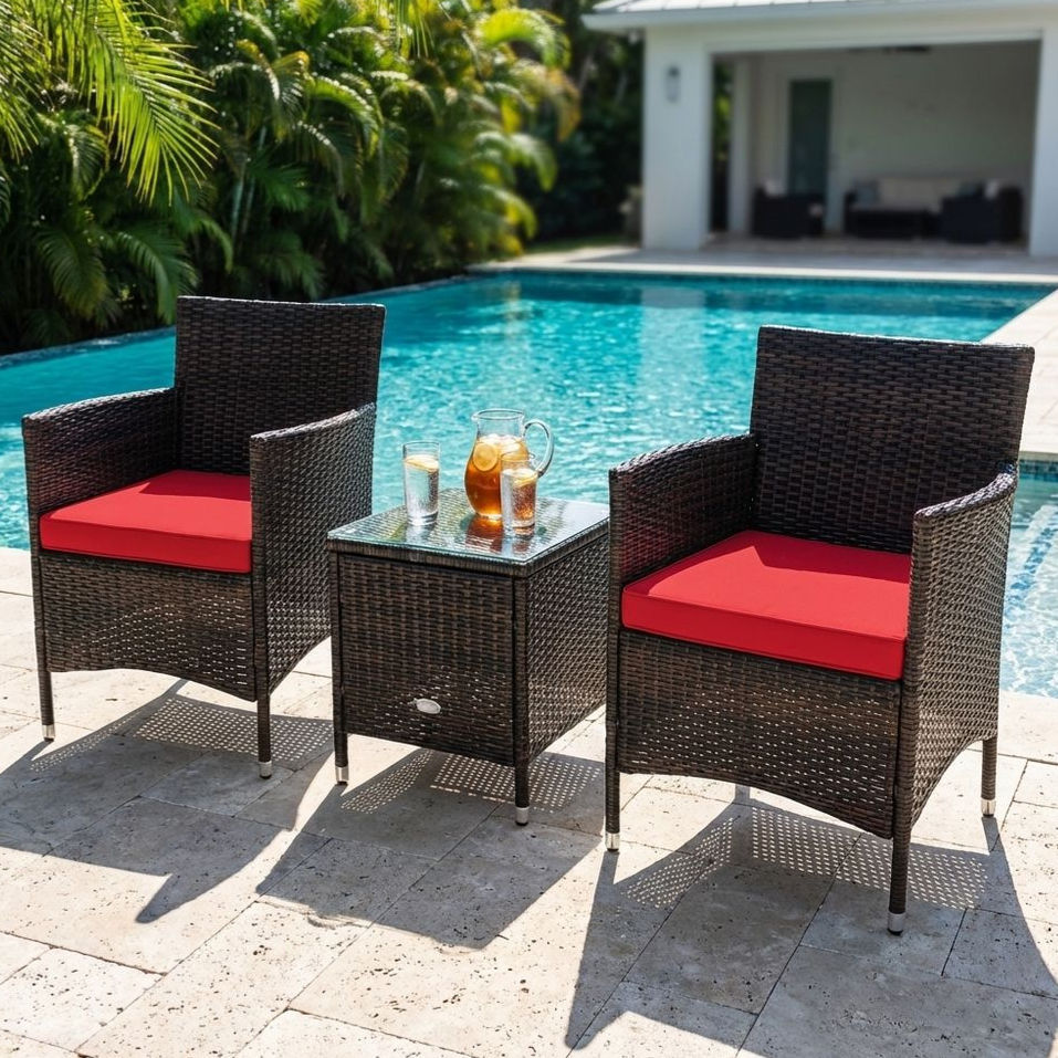 Outdoor wicker patio set with two chairs featuring red cushions and a glass-top table. The setting is by a pool with a pitcher of iced tea and glasses on the table.