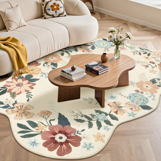 Living room with a beige sofa, floral cushion, wooden coffee table, and a floral-patterned rug with irregular edges. A vase with flowers and books are on the table, and a mustard-yellow throw is draped over the sofa.
