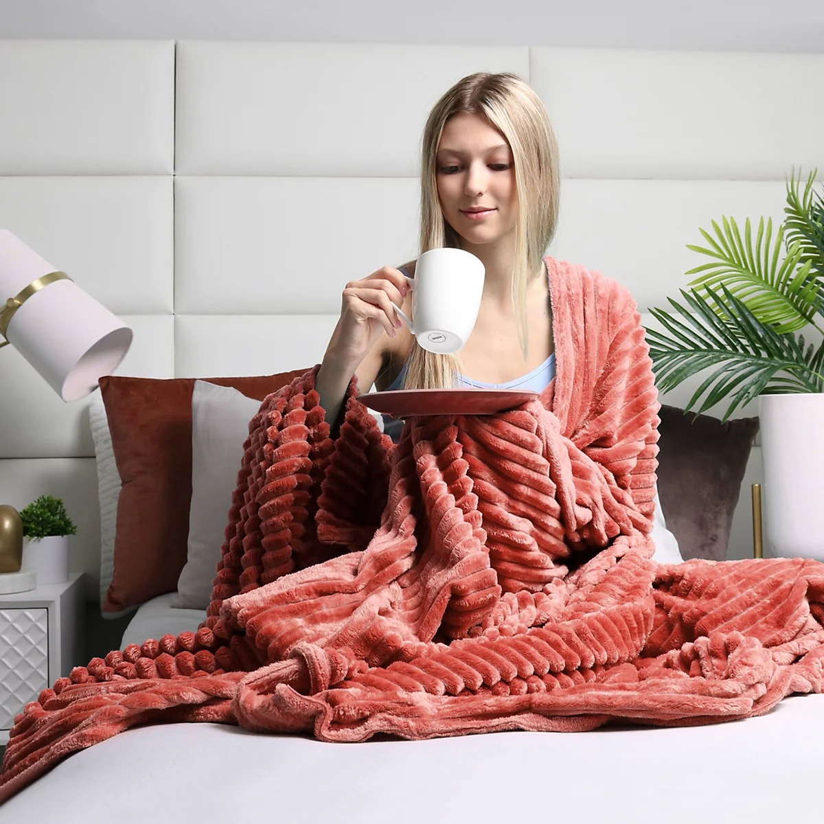 A person sits on a bed wrapped in a textured, plush blanket, holding a white mug on a saucer. The setting includes pillows, a lamp, and a plant in the background.