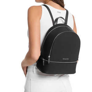 A black Michael Kors backpack with silver zippers is worn by a person in a white sleeveless top and white jeans.