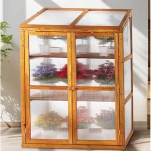 A small wooden greenhouse with a clear polycarbonate paneling has three shelves filled with colorful potted plants. It has a rustic look and two front doors with wooden knobs.