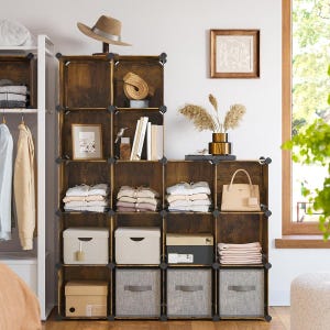 A modular storage shelf with nine cube compartments holds neatly folded clothes, storage bins, a hat, a handbag, and decorative items like books, a frame, and pampas grass in a vase, situated beside a clothing rack and window.