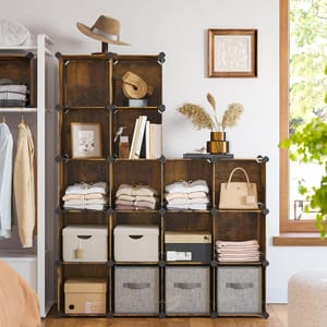 A modular storage shelf with nine cube compartments holds neatly folded clothes, storage bins, a hat, a handbag, and decorative items like books, a frame, and pampas grass in a vase, situated beside a clothing rack and window.