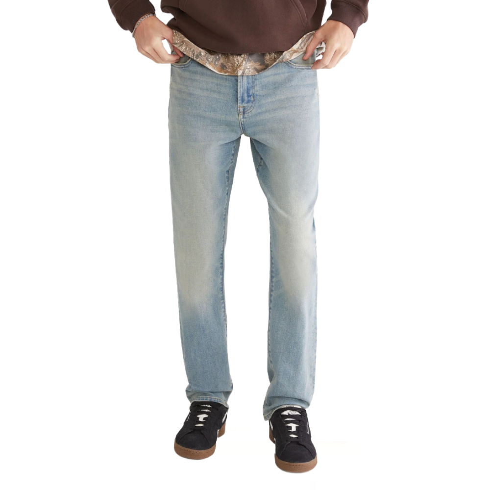 A person wearing light blue jeans and black sneakers with white laces, paired with a brown hoodie. The jeans are being held at the waistband, revealing camouflage-patterned fabric underneath.