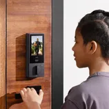 A person is using a digital door lock with a screen displaying a visitor's image. The lock has a sleek design and is mounted on a wooden door.