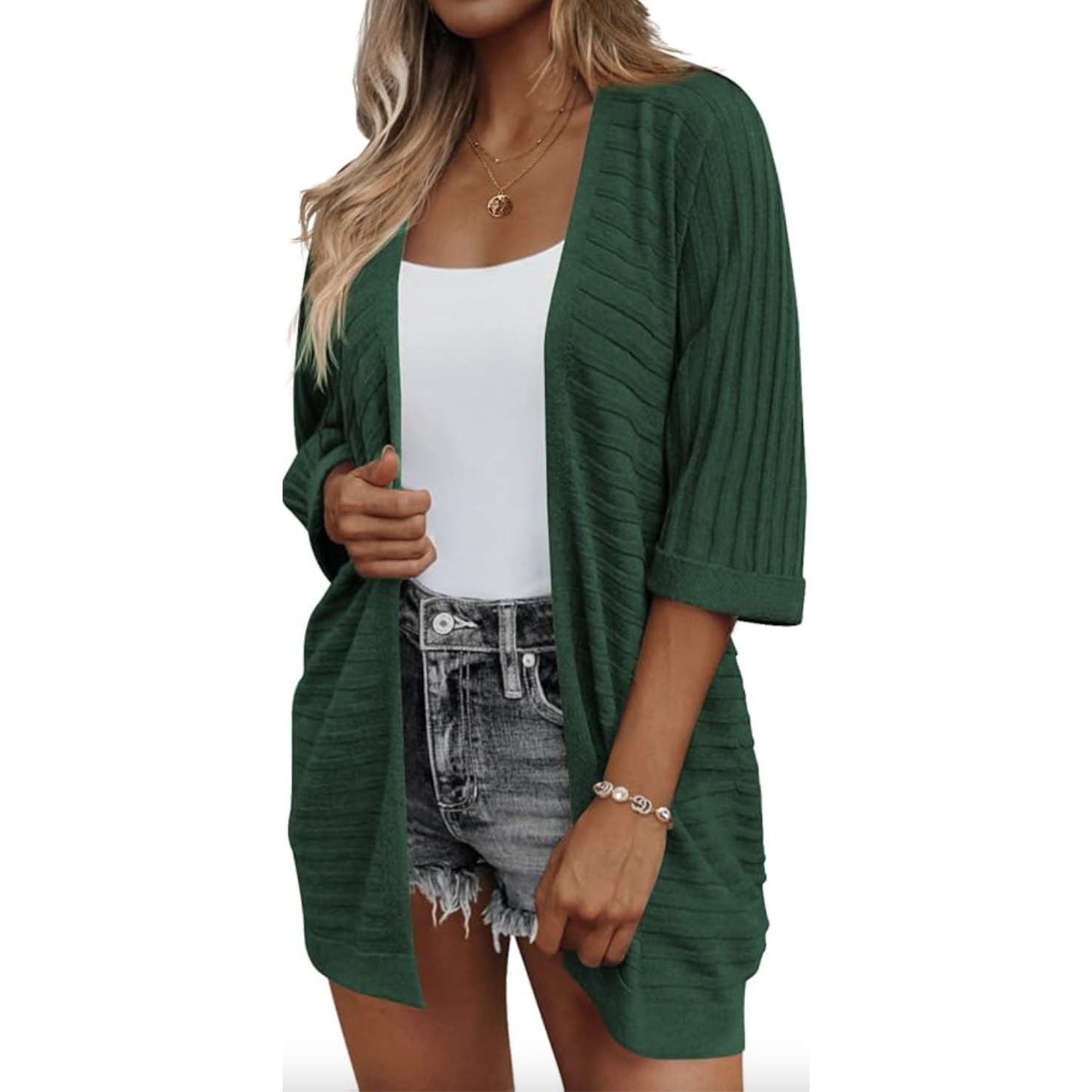A woman is wearing a textured, dark green cardigan with elbow-length sleeves over a white top and high-waisted denim shorts. The cardigan is open-front and ribbed, offering a casual, relaxed fit.