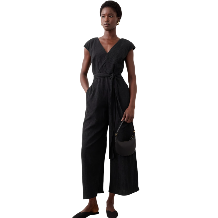 A woman is wearing a black V-neck jumpsuit with short sleeves, a tied waist, and wide legs. She holds a small black handbag and wears black slip-on shoes.