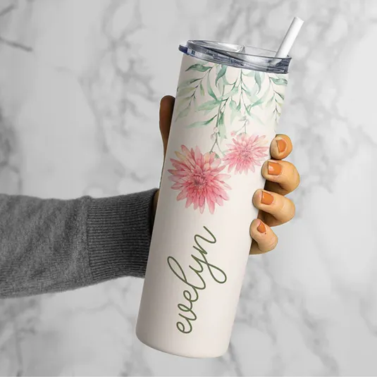 A tumbler with floral design and a name \“Evelyn,\“ featuring a lid and straw, is held by a person with neatly painted nails.