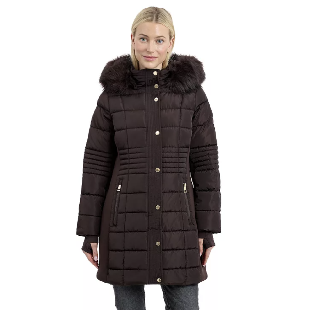 A woman wears a dark brown hooded puffer coat with a faux fur trim and gold-tone button details, paired with dark jeans.