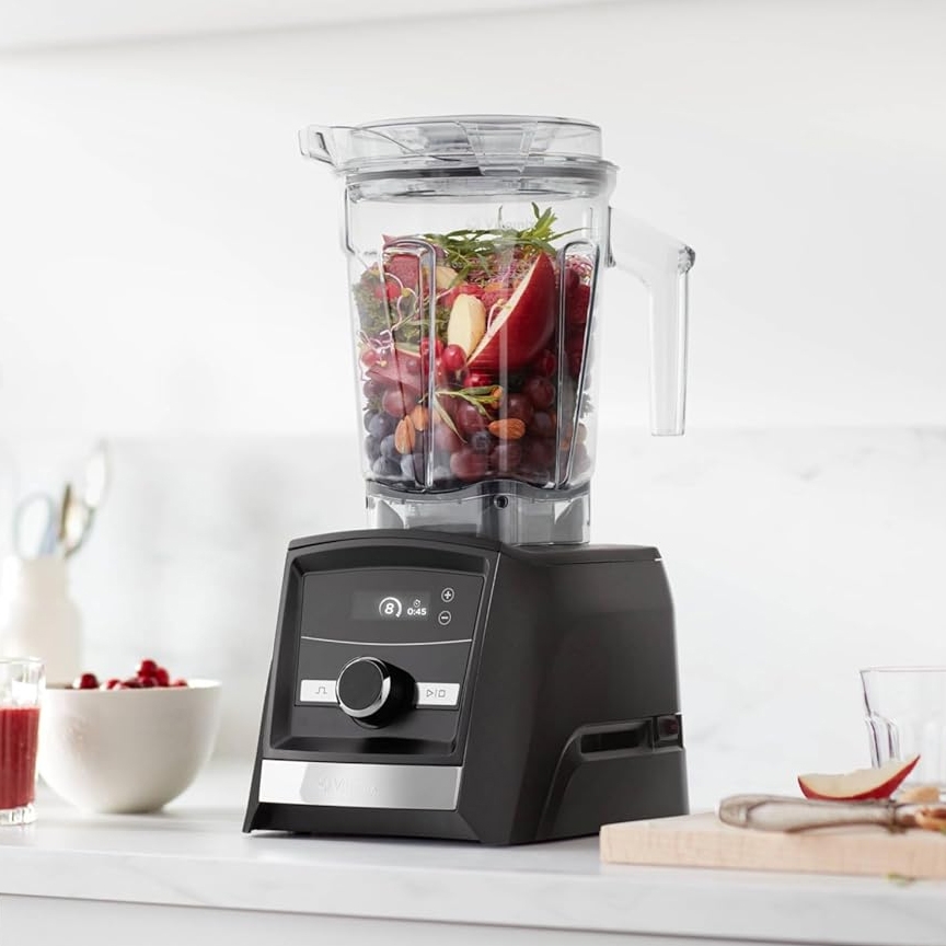 A modern black blender filled with various fruits, including apples and grapes, is displayed on a kitchen counter.