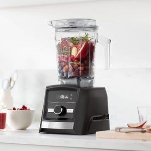 A modern black blender filled with various fruits, including apples and grapes, is displayed on a kitchen counter.
