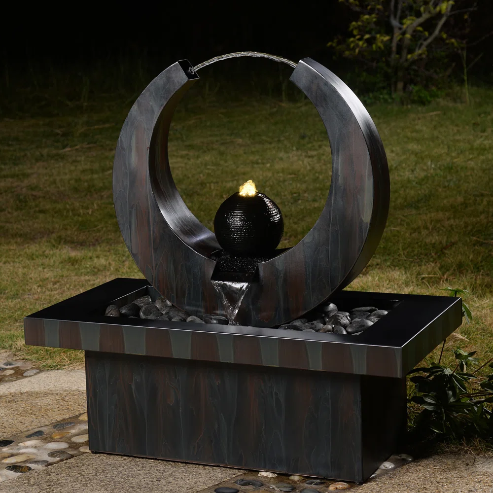 A modern outdoor water feature with a metallic circular frame, a lit central black sphere, and a rectangular base filled with decorative stones, set against a grassy background.