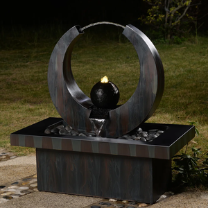 A modern outdoor water feature with a metallic circular frame, a lit central black sphere, and a rectangular base filled with decorative stones, set against a grassy background.