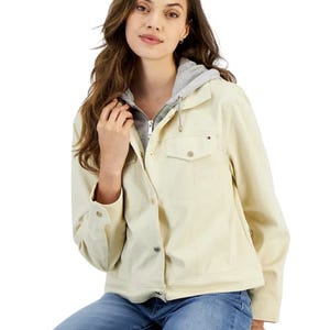 Tommy Hilfiger Women's Hood Track Jacket features a light cream color with a button-up front, snap pockets, and an attached gray hood. It's paired with blue jeans for a casual look.