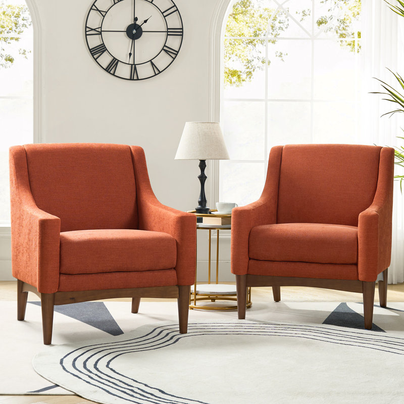 Two mid-century modern armchairs with burnt orange fabric and wooden legs are placed in front of a large window. A round side table with a black lamp sits between them on a light rug with a circular design.