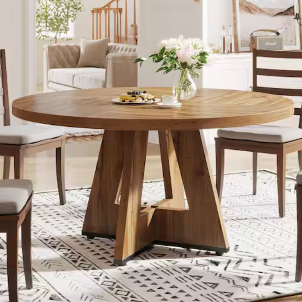 Round wooden dining table with a geometric pedestal base, surrounded by chairs with cushioned seats. A vase with flowers and a plate of snacks are on the table, placed on a patterned rug in a modern dining room setting.
