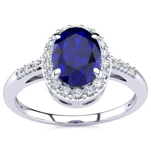 A white gold ring featuring a 1-carat oval blue sapphire center stone, surrounded by a halo of small round diamonds, with additional diamonds set along the band for added sparkle.