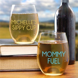 Two wine glasses with humorous text: \“MICHELLE'S SIPPY CUP\“ and \“MOMMY FUEL,\“ next to books and a wine bottle, set against a blurred outdoor background.