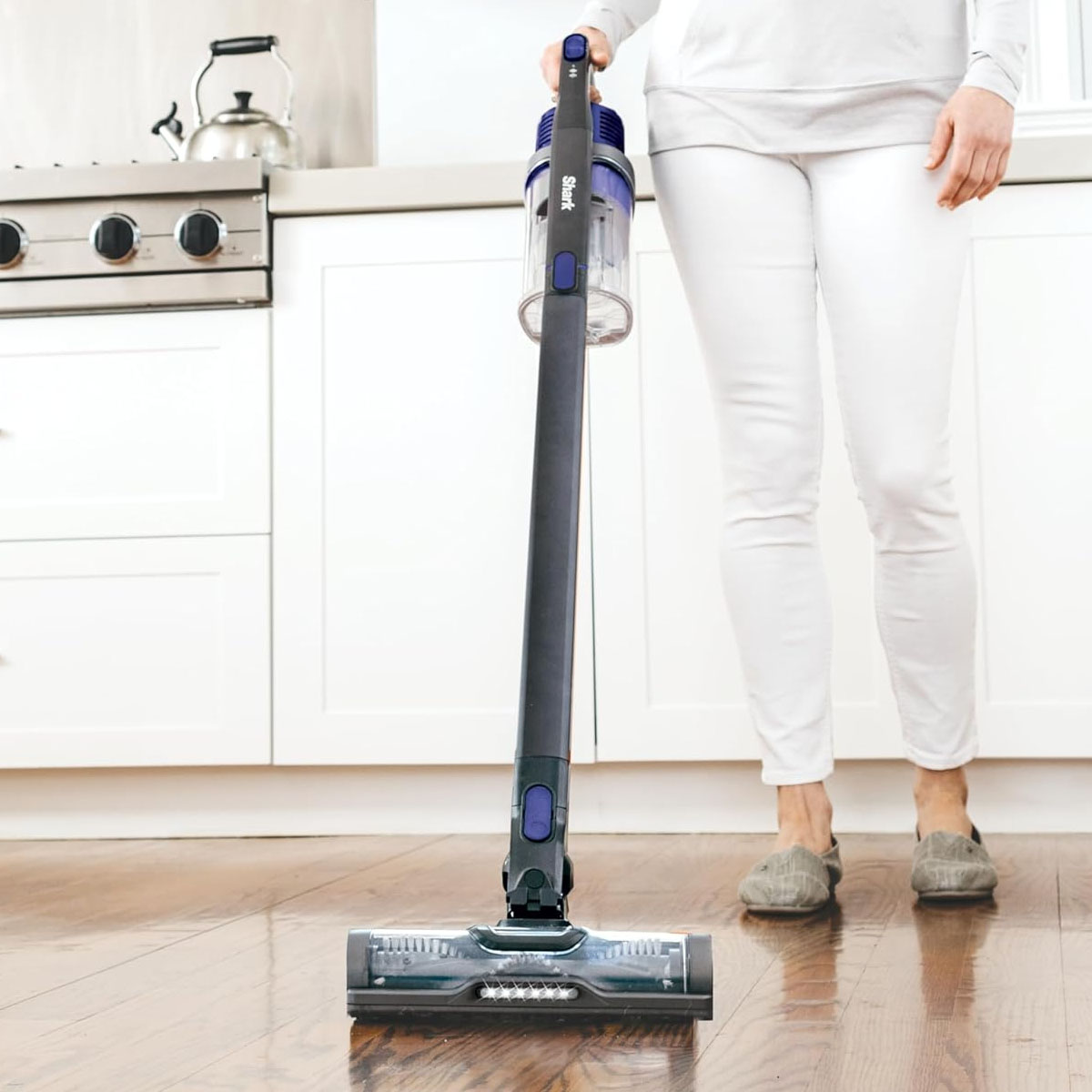 A person is using a Shark cordless stick vacuum to clean a hardwood floor in a kitchen.