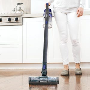 A person is using a Shark cordless stick vacuum to clean a hardwood floor in a kitchen.