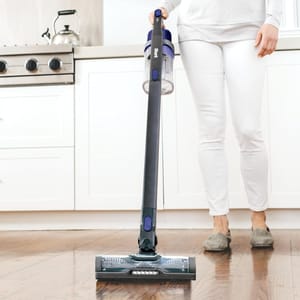 A person is using a Shark cordless stick vacuum to clean a hardwood floor in a kitchen.