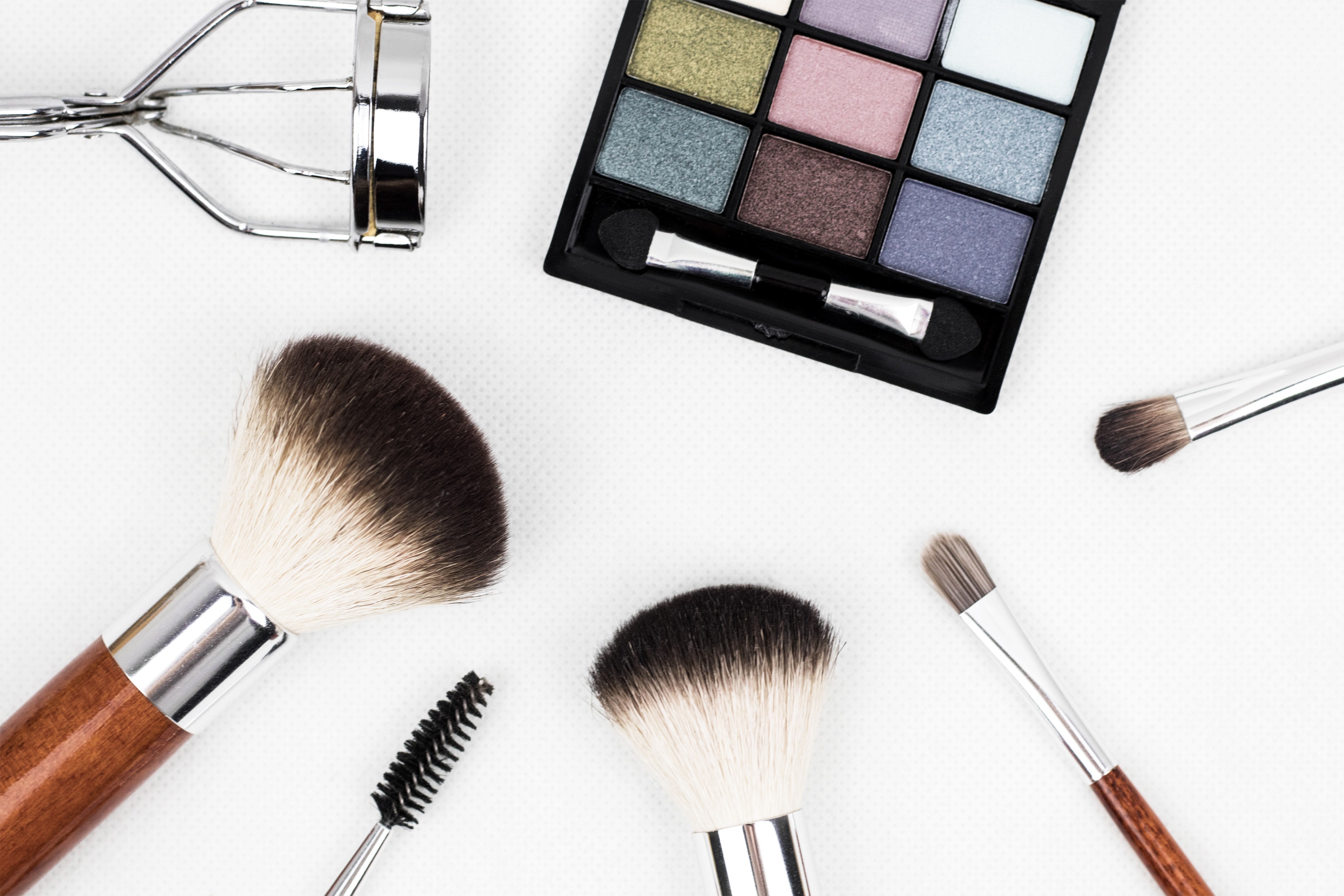 Makeup palette with various shades, eyelash curler, and an assortment of cosmetic brushes on a white background.