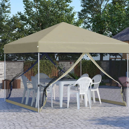 A beige canopy tent with open mesh sides covers a white table and several white plastic chairs, set on a stone patio surrounded by trees and a wooden fence.